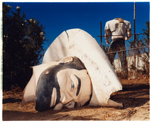 Load image into Gallery viewer, Photograph by Richard Heeps. In the foreground of the photograph is a broken off head of a giant fibreglass sculpture of a cowboy (a Muffler Man). In the background set in a deep blue sky is the muffler man's headless torso.
