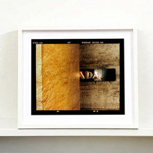 Load image into Gallery viewer, White framed photograph by Richard Heeps. Brown flecked marble walls in different tones. In the middle is half a brown plaque with golden letters showing half an A, followed by a D and an A.