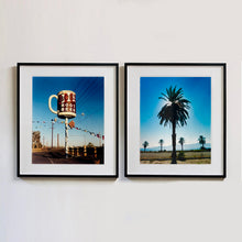 Load image into Gallery viewer, Two black framed photographs by Richard Heeps. A giant model of a mug with Bob's Root Beer written on it sits on top of a giant pole. There is bunting hanging from the pole. It sits alongside a power line on a remote looking American country road. The photograph on the right hand side features a palm tree with palm trees in the background, set in front of a blue sky and the sun filtering from behind the fronds of the tree.