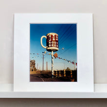 Load image into Gallery viewer, Mounted photograph by Richard Heeps. A giant model of a mug with Bob's Root Beer written on it sits on top of a giant pole. There is bunting hanging from the pole. It sits alongside a power line on a remote looking American country road.