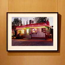 Load image into Gallery viewer, Black framed photograph by Richard Heeps. This photograph depicts a one storey small building &quot;Dot&#39;s Diner&quot; brightly lit with a pink roof, with Hamburgers, Hot Dogs, Shakes, Fries written along the top width of the building.