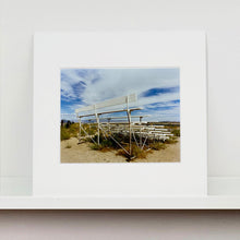 Load image into Gallery viewer, Mounted photograph by Richard Heeps. The back view of a basic white painted grandstand sits in the middle of this photograph. Grass grows up from its base and it sits alone on a sandy ground.