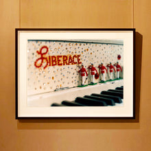 Load image into Gallery viewer, Black framed photograph by Richard Heeps. A close up of Liberace&#39;s piano. This captures the black keys and the fall board which is decorated with crystals and red buttons spelling out Liberace in capitals. There are also 5 red and white metal soldiers positioned marching along the rim.