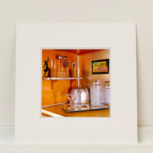 Load image into Gallery viewer, Mounted photograph by Richard Heeps. A drinks corner with martini glasses, cocktail shaker and ice bucket. There is a shelf with cocktail making equipment and it is set against retro orange walls.