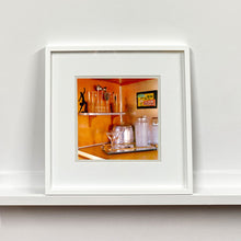 Load image into Gallery viewer, White framed photograph by Richard Heeps. A drinks corner with martini glasses, cocktail shaker and ice bucket. There is a shelf with cocktail making equipment and it is set against retro orange walls.