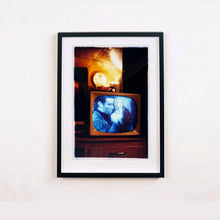 Load image into Gallery viewer, Black framed photograph by Richard Heeps. A retro small brown television sits on a wooden table displaying a black and white film which shows as blue and white. In the background of the television is a warm light.