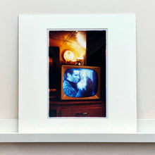Load image into Gallery viewer, Mounted photograph by Richard Heeps. A retro small brown television sits on a wooden table displaying a black and white film which shows as blue and white. In the background of the television is a warm light.