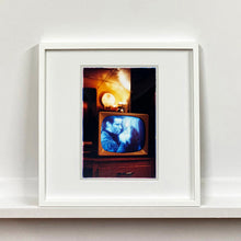 Load image into Gallery viewer, White framed photograph by Richard Heeps. A retro small brown television sits on a wooden table displaying a black and white film which shows as blue and white. In the background of the television is a warm light.