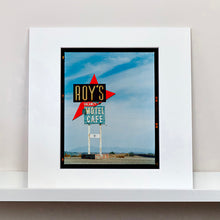 Load image into Gallery viewer, Mounted photograph by Richard Heeps edged by film rebate. A roadside sign on Route 66 in America. The word ROY'S appears in a black sign with a bit red arrow pointing to the left ground, below this VACANCY and on a green square the words MOTEL and CAFE.