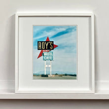 Load image into Gallery viewer, White framed photograph by Richard Heeps. A roadside sign on Route 66 in America. The word ROY'S appears in a black sign with a big red arrow pointing to the left ground, below this VACANCY and on a green square the words MOTEL and CAFE.