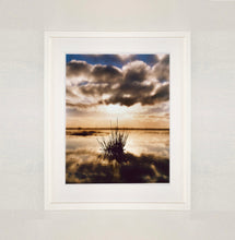 Load image into Gallery viewer, White framed photograph by Richard Heeps. A tussock of grass sits at dusk in fenland water. It is siting under a black and white cloud formation with a golden dusk hue.