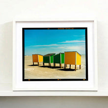 Load image into Gallery viewer, White framed photograph by Richard Heeps. Yellow and green beach lockers stand on their wooden legs on the clear sandy beach.