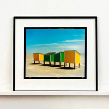 Load image into Gallery viewer, Black framed photograph by Richard Heeps. Yellow and green beach lockers stand on their wooden legs on the clear sandy beach.