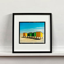 Load image into Gallery viewer, Black framed photograph by Richard Heeps. Yellow and green beach lockers stand on their wooden legs on the clear sandy beach.