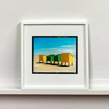 Load image into Gallery viewer, White framed photograph by Richard Heeps. Yellow and green beach lockers stand on their wooden legs on the clear sandy beach.