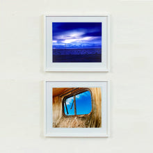 Load image into Gallery viewer, Two white framed photographs by Richard Heeps. The top photograph captures blue light as it hits vast land, mountains and a big sky. The bottom photograph is a blue sky through the window of a dilapidated trailer.