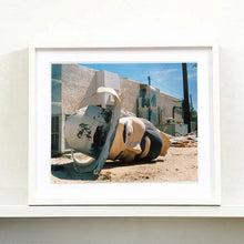 Load image into Gallery viewer, White framed photograph by Richard Heeps. The broken off head of a giant fibreglass sculpture of a cowboy (a Muffler Man) lies on the ground.