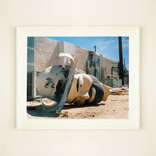 Load image into Gallery viewer, White framed photograph by Richard Heeps. The broken off head of a giant fibreglass sculpture of a cowboy (a Muffler Man) lies on the ground.