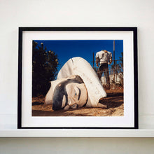 Load image into Gallery viewer, Black framed photograph by Richard Heeps. In the foreground of the photograph is a broken off head of a giant fibreglass sculpture of a cowboy (a Muffler Man). In the background set in a deep blue sky is the muffler man's headless torso.