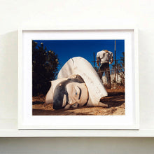 Load image into Gallery viewer, White framed photograph by Richard Heeps. In the foreground of the photograph is a broken off head of a giant fibreglass sculpture of a cowboy (a Muffler Man). In the background set in a deep blue sky is the muffler man's headless torso.