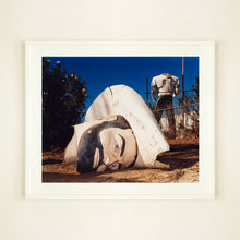 Load image into Gallery viewer, White framed photograph by Richard Heeps. In the foreground of the photograph is a broken off head of a giant fibreglass sculpture of a cowboy (a Muffler Man). In the background set in a deep blue sky is the muffler man's headless torso.