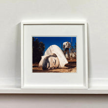 Load image into Gallery viewer, White framed photograph by Richard Heeps. In the foreground of the photograph is a broken off head of a giant fibreglass sculpture of a cowboy (a Muffler Man). In the background set in a deep blue sky is the muffler man's headless torso.