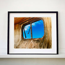 Load image into Gallery viewer, Black framed photograph by Richard Heeps. The interior of an eroded trailer, looking through the broken window to a deep blue sky.