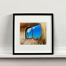 Load image into Gallery viewer, Black framed photograph by Richard Heeps. The interior of an eroded trailer, looking through the broken window to a deep blue sky.