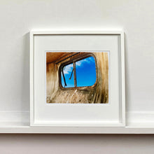 Load image into Gallery viewer, White framed photograph by Richard Heeps. The interior of an eroded trailer, looking through the broken window to a deep blue sky.