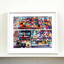 Load image into Gallery viewer, White framed photograph by Richard Heeps. Three compartmentalised shop shelves, each compartment has coloured plastic dice, counters, circular containers, cards, poker chips, and other plastic type games items.