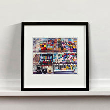 Load image into Gallery viewer, Black framed photograph by Richard Heeps. Three compartmentalised shop shelves, each compartment has coloured plastic dice, counters, circular containers, cards, poker chips, and other plastic type games items.