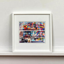 Load image into Gallery viewer, White framed photograph by Richard Heeps. Three compartmentalised shop shelves, each compartment has coloured plastic dice, counters, circular containers, cards, poker chips, and other plastic type games items.