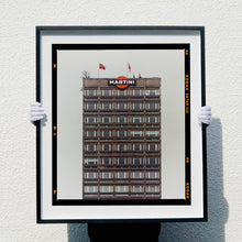 Load image into Gallery viewer, Black framed photograph held by photographer Richard Heeps. High rise offices with Martini logo on the top facade.
