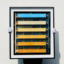 Load image into Gallery viewer, |A black framed photograph held by photographer Richard Heeps. Photograph of an apartment building with coloured balconies, blue at the top balcony and then fading from light yellow to a sunburnt yellow at the bottom.
