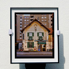 Load image into Gallery viewer, Black framed photograph held by photographer Richard Heeps. A traditional Italian Tobacconist shop, here in a Swiss Cottage style building set against a vast urban apartment block.