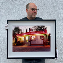 Load image into Gallery viewer, Black framed photograph held by Richard Heeps. This photograph depicts a one storey small building "Dot's Diner" brightly lit with a pink roof, with Hamburgers, Hot Dogs, Shakes, Fries written along the top width of the building.