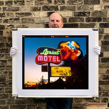 Load image into Gallery viewer, Photograph held by photographer Richard Heeps. The photograph is taken in the evening, a neon sign belonging to the Lariat Motel, with a cowboy on horseback with a lasso on the right of the words, half way down the sign post is the yellow sign God Bless America.