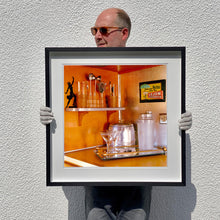 Load image into Gallery viewer, Black framed photograph held by photographer Richard Heeps. A drinks corner with martini glasses, cocktail shaker and ice bucket. There is a shelf with cocktail making equipment and it is set against retro orange walls.