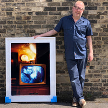 Load image into Gallery viewer, Photograph held by photographer Richard Heeps. A retro small brown television sits on a wooden table displaying a black and white film which shows as blue and white. In the background of the television is a warm light.