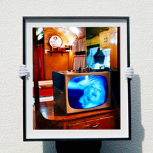 Load image into Gallery viewer, Black framed photograph held by photographer Richard Heeps. A retro small brown television sits on a wooden table displaying a black and white film, with the face of Marilyn Monroe filling the screen, it shows as blue and white. In the background of the television is a warm wood-panelled light.