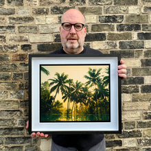 Load image into Gallery viewer, Black framed photograph held by photographer Richard Heeps. Green palms trees against a warm golden sky, and reflected in a pool below.