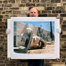 Load image into Gallery viewer, Photograph held by photographer Richard Heeps. The broken off head of a giant fibreglass sculpture of a cowboy (a Muffler Man) lies on the ground.