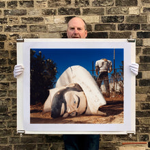 Load image into Gallery viewer, Photograph held by photographer Richard Heeps. In the foreground of the photograph is a broken off head of a giant fibreglass sculpture of a cowboy (a Muffler Man). In the background set in a deep blue sky is the muffler man's headless torso.