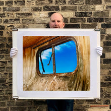 Load image into Gallery viewer, Photograph held by photographer Richard Heeps. The interior of an eroded trailer, looking through the broken window to a deep blue sky.