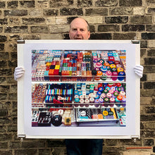 Load image into Gallery viewer, Photograph held by photographer Richard Heeps. Three compartmentalised shop shelves, each compartment has coloured plastic dice, counters, circular containers, cards, poker chips, and other plastic type games items.