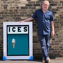 Load image into Gallery viewer, White framed photograph held by photographer Richard Heeps.  At the top black letters spell out ICES and below is depicted a 99 icecream cone sitting left of centre against a blue teal coloured background.