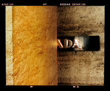 Load image into Gallery viewer, Photograph by Richard Heeps. Brown flecked marble walls in different tones. In the middle is half a brown plaque with golden letters showing half an A, followed by a D and an A.