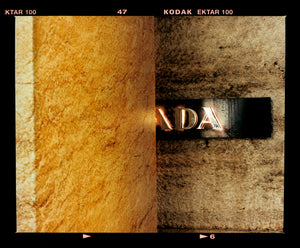 Photograph by Richard Heeps. Brown flecked marble walls in different tones. In the middle is half a brown plaque with golden letters showing half an A, followed by a D and an A.