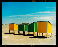 Load image into Gallery viewer, Photograph by Richard Heeps. Yellow and green beach lockers stand on their wooden legs on the clear sandy beach.