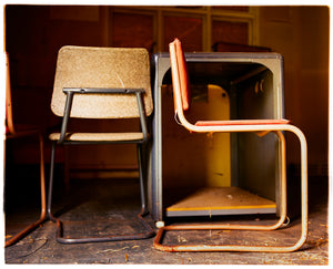 Photograph by Richard Heeps.  Two metal framed chairs with padded seats and backs. On the left hand side of the photo is one of the chairs facing backwards, on the right handside is a chair facing right. The chairs sit next to a metal cabinet. The floor is covered in small shavings of debris.